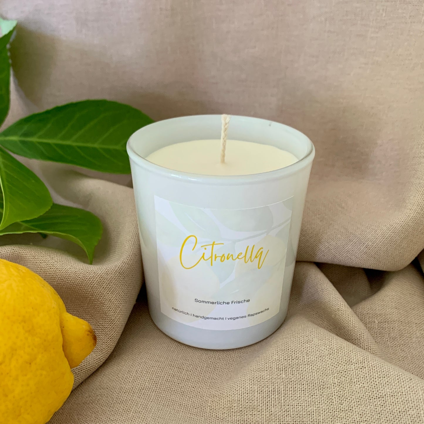 Citronella Kerze aus Rapswachs in einem weißen Glas. Dekoriert auf einem beigen Stoff mit einer Zitrone und einem Blätterzweig drumherum.