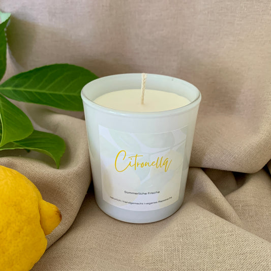 Citronella Kerze aus Rapswachs in einem weißen Glas. Dekoriert auf einem beigen Stoff mit einer Zitrone und einem Blätterzweig drumherum.
